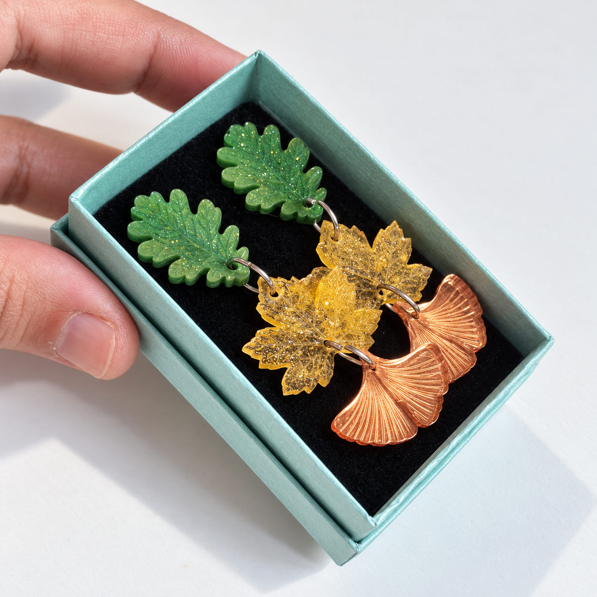 Pendientes Danza de Hojas de Sergi Bastian: hojas verde nacarado, amarillo glitter y ginkgo espejo cobre en caja turquesa.