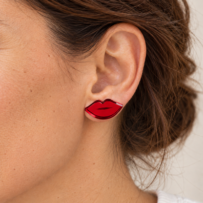 Mini Labios en rojo espejo colocados como pendientes de botón sobre el lóbulo, con la oreja visible en primer plano.