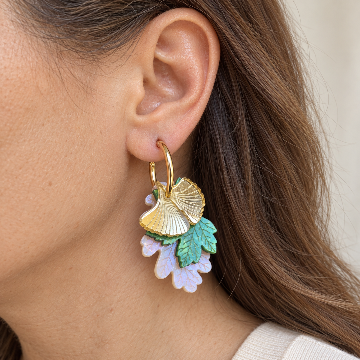 Pendiente Luz de Ginkgo puesto en una oreja, con aro dorado y tres hojas superpuestas; primer plano sin mostrar el rostro.